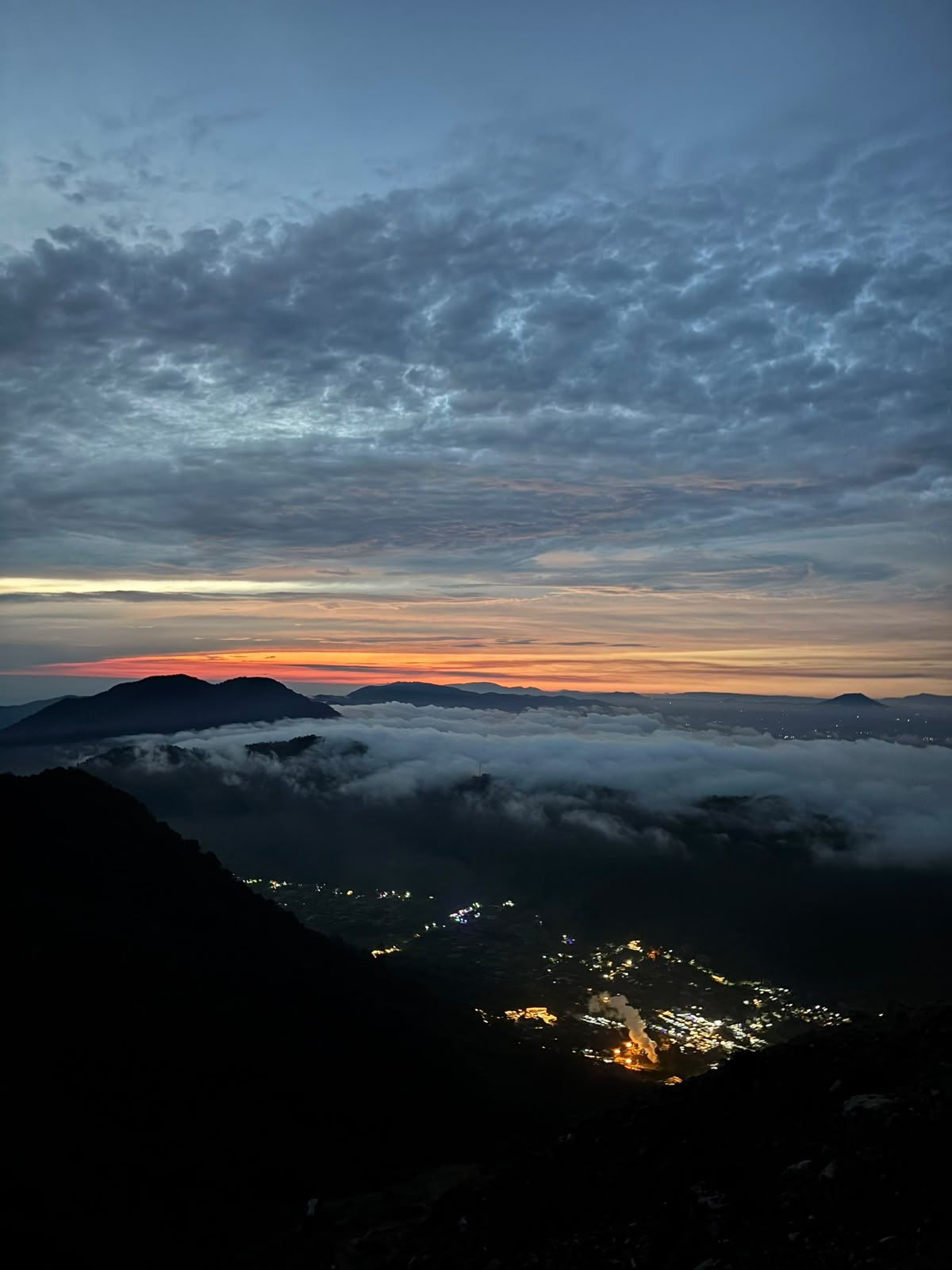 Dawn above the clouds. First light on the horizon, with village lights still visible in the valley.